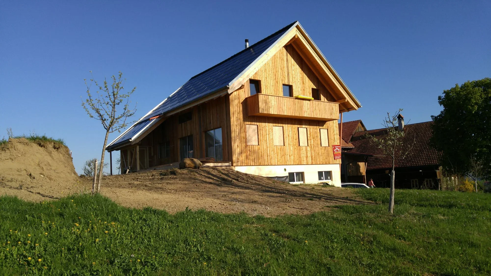 Neues Holzhaus mit Satteldach und Balkon auf einem frisch angelegten Hügel unter blauem Himmel, rechts das alte Bauernhaus