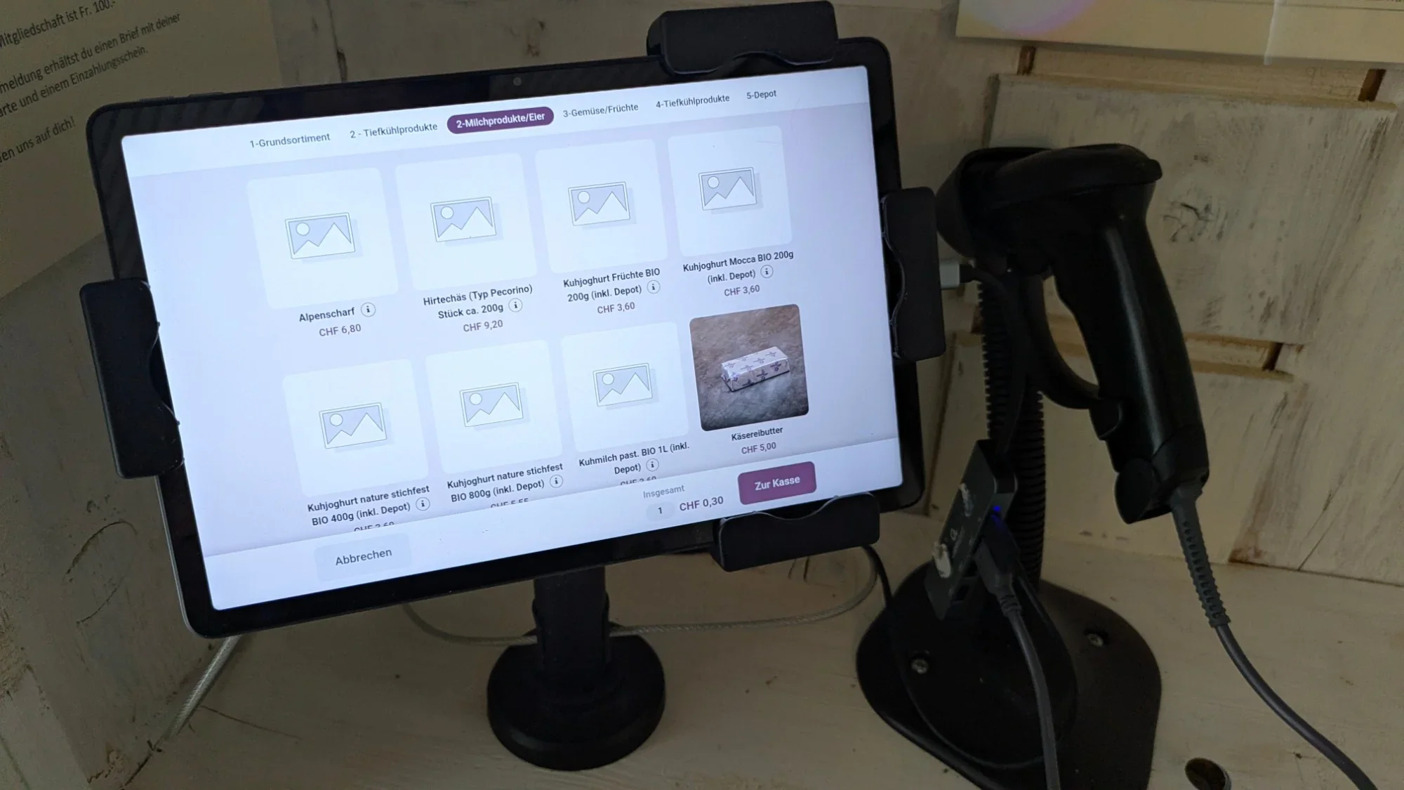 Tablet mit Scanner am Kassenplatz, Bildschirm zeigt Milchprodukte und Eier mit Preisen, darunter Alpenscharf und Hirtechäs