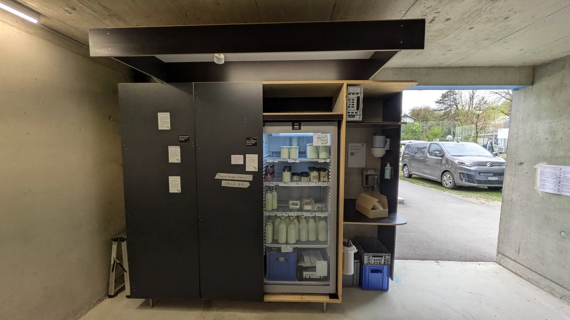 Schwarzer Schrank mit Kühlschrank, Regalen und Milchharassen im Vorraum der Velogarage