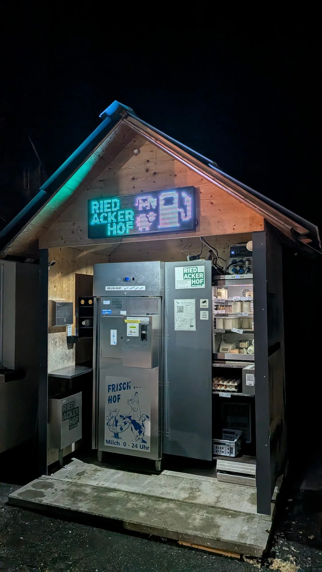Beleuchtetes Holzhäuschen mit Milchautomat, Kühlschrank mit Joghurt, Käse und Eiern bei Nacht